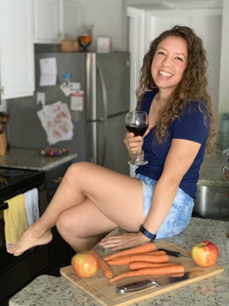 Coach Kristin holding a glass of wine near a cutting board with prepared vegetables.